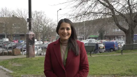 Local Democracy Reporting Service Shows a woman with dark hair, wearing a red coat smiling at the camera. In the distance are some shops and benches, and a tall tree.