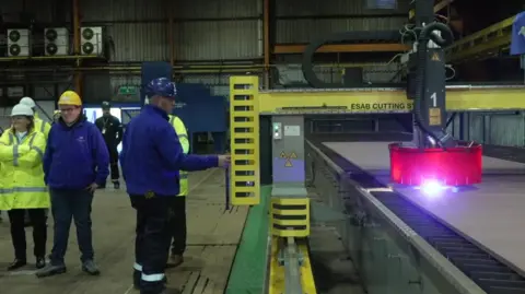 BBC A man in a blue uniform is seen cutting a piece of steel. 