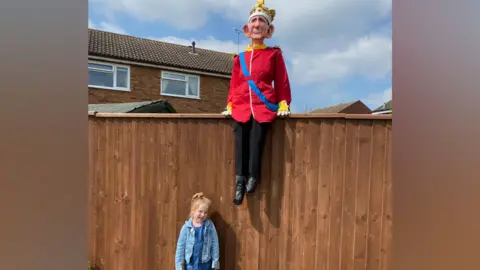 Contributed A little girl stands in front of a tall wooden fence. She has blonde hair and wears a denim jacket, blue dress, white tights and white trainers. A sculpture of King Charles sits on top of the fence wearing a crown, red regal coat, black trousers and black shoes.