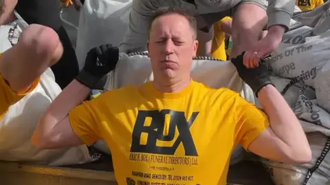 A man wearing a bright yellow event T-shirt and black gloves stands with his eyes closed and a strained expression as a large white sack of coal is positioned on to his shoulders. Someone behind him, dressed in a grey hoodie, is helping lift the sack. Around them are other competitors and sacks of coal.