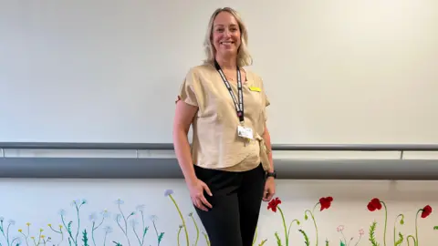 BBC Consultant Nurse Emma Clark stands in the new Peddars Way End of Life Unit at the Queen Elizabeth Hospital in King's Lynn. She is wearing a beige coloured top and black trousers. The bottom half of the walls are subtly painted with daisy's, buttercups and poppies to make the hospital environment seem less clinical. 