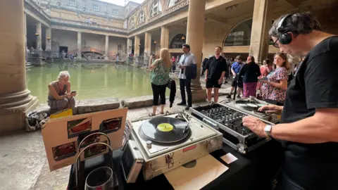 A man with black headphones on is spinning records at two turntables in the foreground. People can be seen milling around in front of him and around the stone courtyard surrounding the thermal bath.