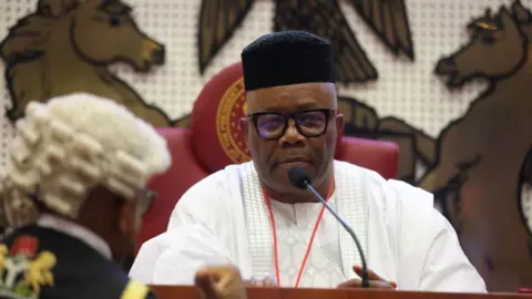 AFP Godswill Akpabio sits on a red seat in front of a microphone. He wears black-rimmed glasses, a black hat and a white shirt