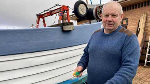 Fiona Irving / BBC A white man with short white hair looks at the camera. He is wearing a blue jumper with patches on the shoulders. In his hand is a green fishing line. He is standing next to a white and blue boat.