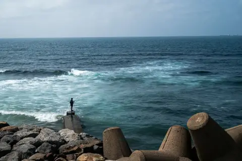 JEWEL SAMAD / AFP / GETTY IMAGES A man fishes from the shore. Behind him are large rocks and concrete wave breakers.