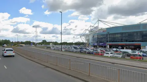 Google Google Streetview image of Judds Lane, in Coventry. Two lanes run in each direction, with a large number of cars in the background, parked in the stadium car park. The stadium itself is on the right
