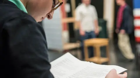 A girl with glasses wearing a black jumper and a green t-shirt holding a script. The view is from over her shoulder and she is looking down at the script. The background is blurred and shows two people standing behind a chair. 