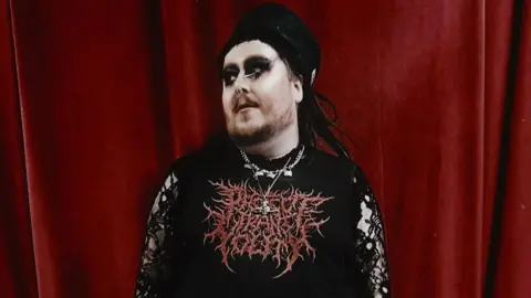 DJ Mathu A person with gothic make-up, several chains around their neck and a top with horror-style red lettering stands in front of a blood-red curtain, looking off to the side.