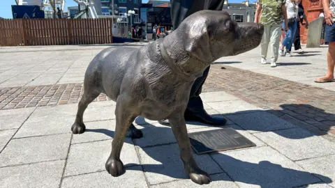 A bronze statue of a dog on the pavement beside the statue of Rhodri. He looks like a Labrador with a collar and it is looking up with his head turned towards Rhodri.