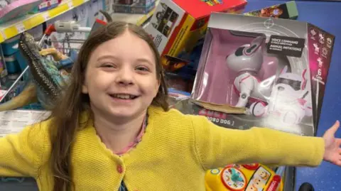 Family photograph Ivy in a yellow knitted cardigan with toys in the background. Her arms are outstretched and she smiles widely. 