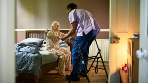 Young male care worker helping woman off bed with walking frame nearby - stock photo.