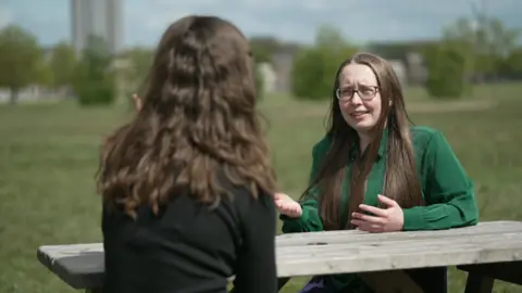 Two women with brown hair sitting on a park bench facing each other having a discussion. One woman is wearing a black top and the other is wearing a green top