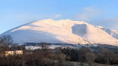 Skiddaw mountain is covered in snow. It is a bright sunny day. The trees in the foreground are bare and have not been hit by the snow.
