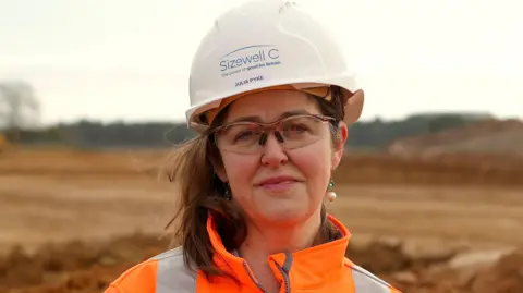 Martin Giles/BBC Julia Pyke smiles at the camera while standing within the Sizewell C construction site. She wears a white hard helmet and orange hi-vis jacket. She has short brown hair and wears clear safety goggles. 