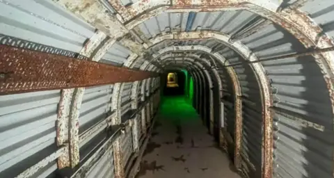 Sussex Trail Events An underground tunnel is lined with corrugated metal and has a concrete floor. An eerie green light can be seen towards the far end