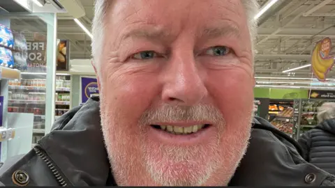 A selfie of Dave Turner. He has a short grey beard and moustache, and grey hair. He's standing in a super market with the fruit and veg aisle in the background.