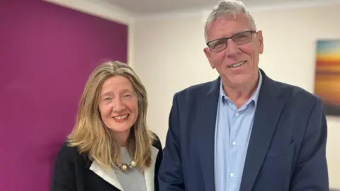 Jon Wright/BBC Ali Oxborough and Brian Tobin stand next to each other inside an office with a purple wall in the background