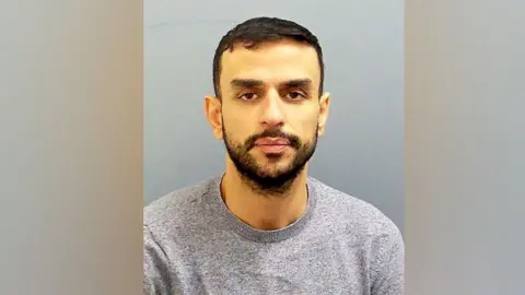 SBNA A custody photo of Mohammed Altaher, who has short black hair and a close-cropped black beard. He is wearing a grey sweatshirt. 