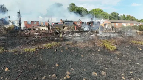 Tim Bourne burned out grass in the foreground with smoke billowing from roofless black bungalows damaged in the fire 