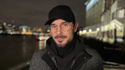Charlie Rose/BBC After dark, standing next to the River Thames on London's South Bank, a bearded Shane Manogue, in glasses, black hoodie and baseball cap, looks at the camera lens.