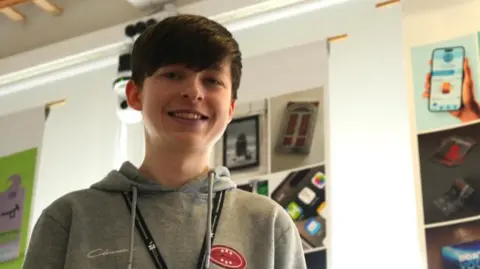 A man with dark hair in a grey top is wearing a lanyard around his neck. A number of images are on the wall behind him.