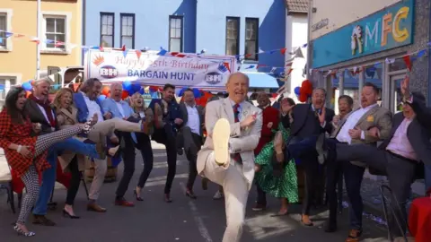 Presenters and experts stand in a semi-circle doing the Bargain Hunt kick. The team are stood in a street under a Happy 25th Birthday Bargain Hunt banner. 