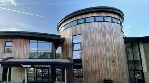 The exterior of the Roundhouse, it has a modern design with a round central building with wooden panelling. 