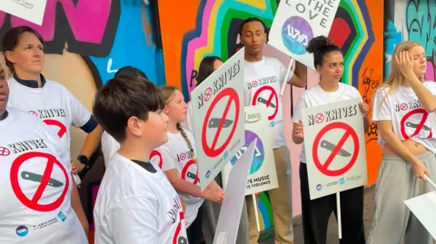 Wycombe Youth Action A group of people wearing matching white anti-knife-crime T-shirts stand beneath a brightly coloured graffiti mural in a covered public walkway. Several members of the group hold placards displaying a red crossed-out knife symbol and the words “NO KNIVES”. One placard reads “WHERE IS THE LOVE” with a circular logo beneath the text. The group is gathered along the wall, with some signs partially overlapping.