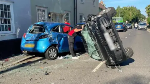 The aftermath of a crash on West Street in Coggeshall. A black car is on its side having turned over. Its windscreen is smashed and there is damage to the front of the car. There is a light blue vehicle which has extensive damage to its rear and side following the crash. There is glass and broken parts of the cars on the ground, and there are two people stood around the crashed vehicles.