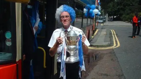 Supplied A person stands beside a bus decorated with blue and white balloons, holding a large silver trophy with ribbons.