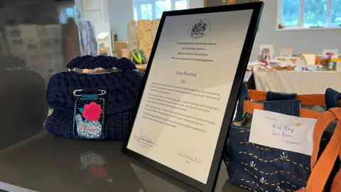 Tom Jackson/BBC A framed King's Award for Voluntary Service certificate perched on a table in between two handmade bags. 