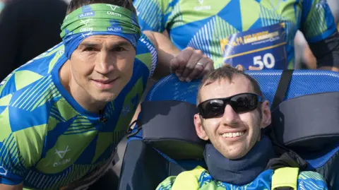 PA Media Kevin Sinfield wears a blue and green shirt and matching headband as he leans close to Rob Burrow, who is wearing the same top and sunglasses as he sits in a wheelchair. Both men are smiling and looking towards the camera.