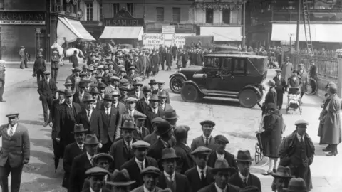 Topical Press Agency / Getty A queue comprising more than 100 men, all dressed smartly with hats, winds through a city centre square. 