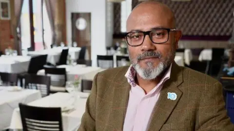 A man with a shaven head, in glasses, standing in a restaurant