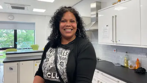 Evelyn with shoulder length black hair in tight curls wearing a long sleeve black top with white floral design in a big square on the front. She is standing in a community kitchen smiling at the camera.