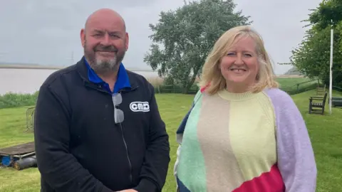 Sally Fairfax / BBC A man in a black zip-up tracksuit top is smiling at the camera next to a blonde woman in a geometric, colourful jumper, who is also smiling. They are both standing on the grassy banks of a river.