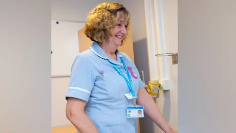 Willen Hospice Nurse Carole Marley wearing her pale blue short sleeved nurse's uniform in a bedroom at the hospice. She is looking down to one side and smiling/chatting with the patient, who is off camera. She has curly golden brown hair and wears a bright blue lanyard with her ID pass.