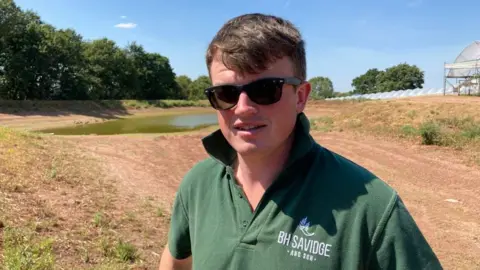 Alistair Binney / BBC There is a man with short brown hair. It is brushed over in a fringe. He has black sunglasses on and a dark green polo shirt with all of his buttons undone. On the right breast pocket area, it has his company name 'BH Savidge and Son.' Behind him is his reservoir storage which has mostly been used up.