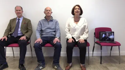 The Schuman family, sat on red hospital chairs next to each other. Christopher, Jeremy, Susan and Philip (on a laptop screen joined virtually)