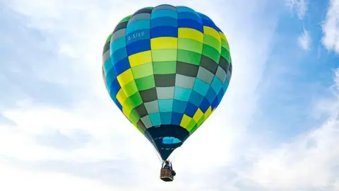 Claire Hartley A solitary multi-coloured hot-air balloon hovers in the air. A partly-cloud covered blue sky can be seen behind it.