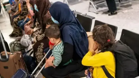 A group of refugees - predominately made up of women and children - sit on chairs in what looks like an airport. They have suitcases around them. Some are carrying young children while other children sit in the seats.