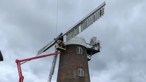 Sue Baxter Under a dark grey sky, the top of the windmill, which is a round red brick building with a white dome on top, has a cherry picker lifting men to the white sails as they remove one