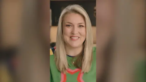 Sara Davies is smiling at the camera. She has long blonde hair and wears a green jumper