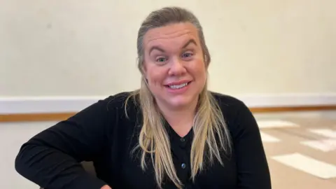 Amber Regnard pictured with long straight blonde hair. She is smiling at the camera and wearing a black cardigan. 