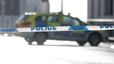 Blue and white police tape. An out of focus police car is in the background.