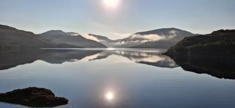 Andrew Beattie A mountain range, which is reflected on a loch, with the sun high in the picture