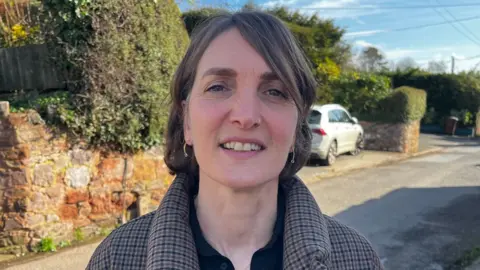 A woman with a nose ring and silver ear rings is standing in a sunny street in a town in Devon. She is wearing a brown checked coat with a black shirt underneath. 