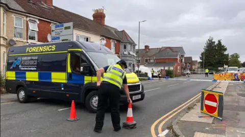 Police van which says 'forensics' with a police officer laying out cones.