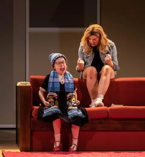 Ste Murray A production image of the play where an actress playing a child sits on a sofa wearing a Coventry City scarf and hat. Next to her an actress playing an adult sits on the back of the red sofa. 