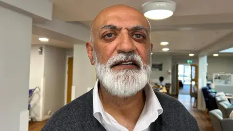 Mahesh looks at the camera. He is bald, with a trimmed white beard, and is wearing a white shift and a grey jumper. A care home lounge with armchairs can be seen in the background. 